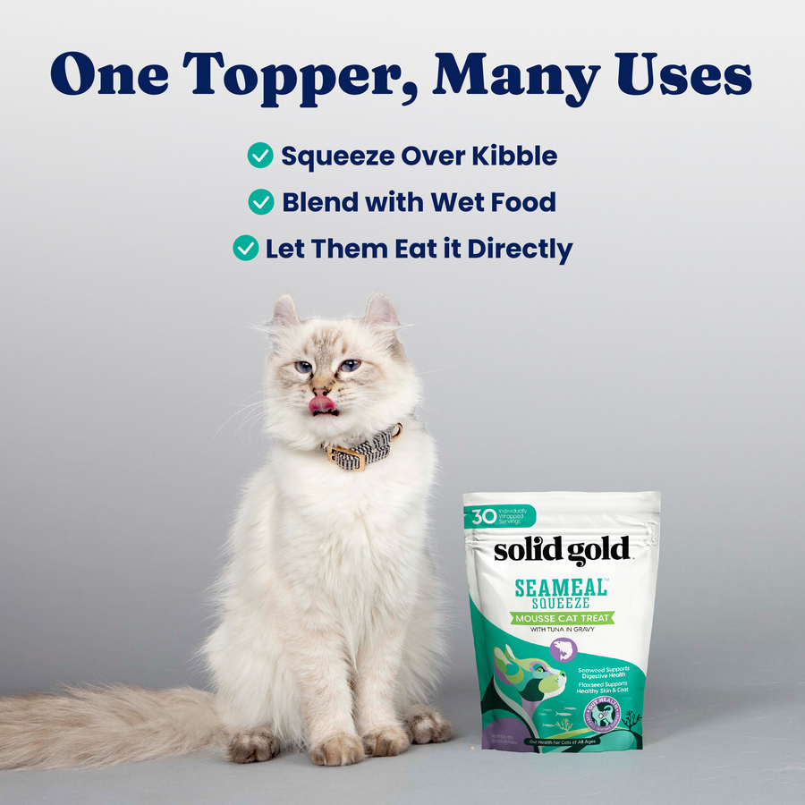 A fluffy white cat sits next to a pouch of SeaMeal Squeeze Mousse Treat, licking its lips.